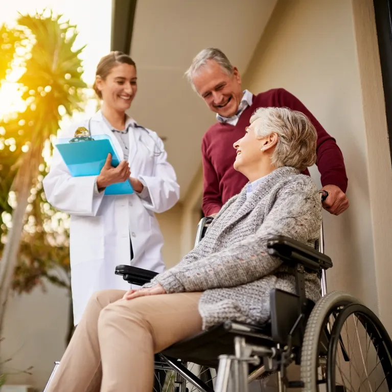 A caregiver holding a clipboard while smiling at an elderly woman in a wheelchair, with her family member standing beside her, creating a warm, supportive moment.