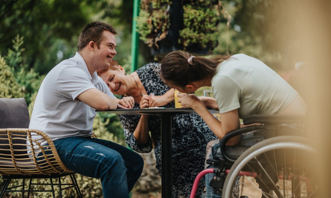 Three friends laughing and enjoying each other’s company at a table outdoors, with one in a wheelchair, creating a joyful and inclusive moment.
