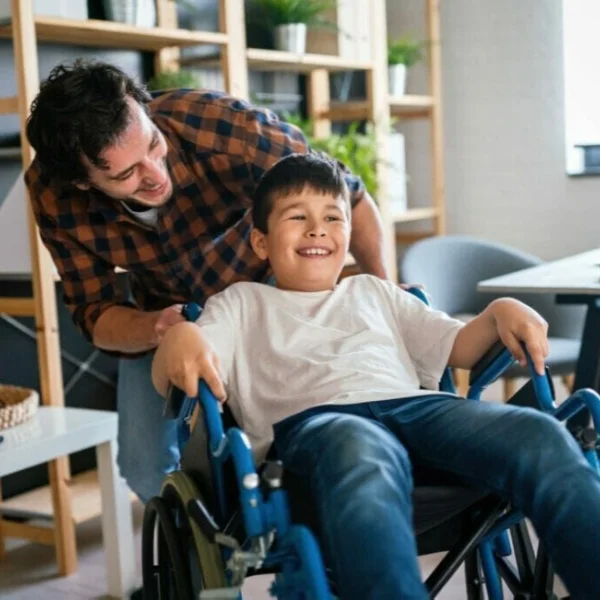 A caregiver pushing a smiling young boy in a wheelchair, both sharing a joyful moment in a bright, cozy indoor space.